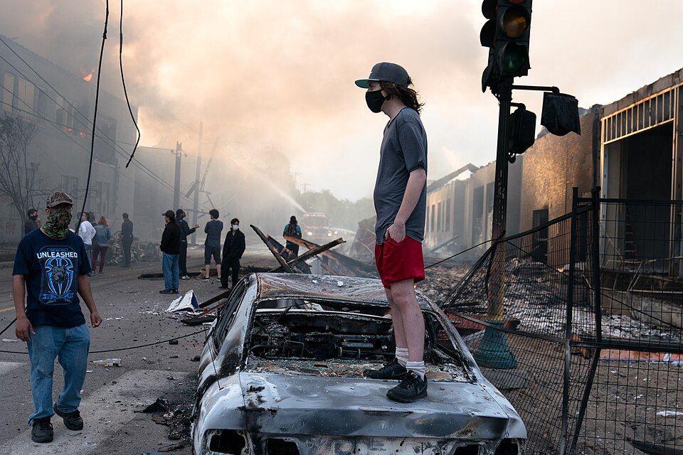 During the morning hours on May 28, 2020, a man stands casually looking northward on a burned out car as several buildings burn and smolder behind him on East 29th Street, near the intersection of South 26th Avenue, in Minneapolis, Minnesota, United States. The fires were caused by acts of arson dur