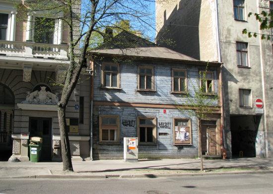 Stabu Street, 18B (Riga)
Location: Latvia, Riga
Coordinates: 56.9573614, 24.1254547

Caption: A weathered, light-blue wooden house stands on a city street, flanked by older buildings. Its windows are framed in dark wood, with graffiti visible on the facade. A small, ornate stone archway and a green 