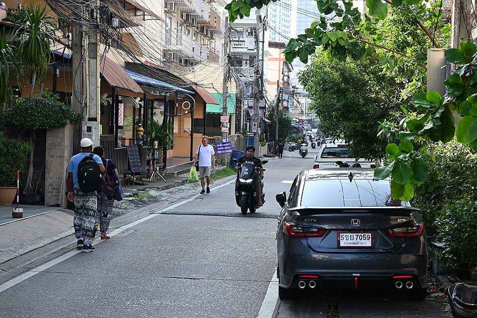 This photo depicts a busy city street with a man riding a motorcycle down the middle of the road. There are several cars parked on the side of the street, and a few people are walking along the sidewalk. Some of the pedestrians are carrying backpacks and handbags.
In addition to the motorcycle, ther