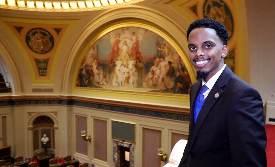 "First-term legislator Omar Fateh shows off the Minnesota Senate chamber in the state Capitol in St. Paul. Behind him is the mural "Minnesota: Granary of the World.""