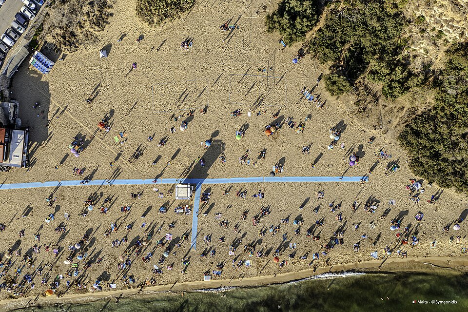 A vertical view of Golden Bay in Malta, focusing on the Il-Lippija area. The beach is filled with people swimming in the clear waters and sunbathing on the golden sand. Umbrellas dot the landscape, casting shadows over sunbathers, while the bright sun enhances the natural beauty of the area.