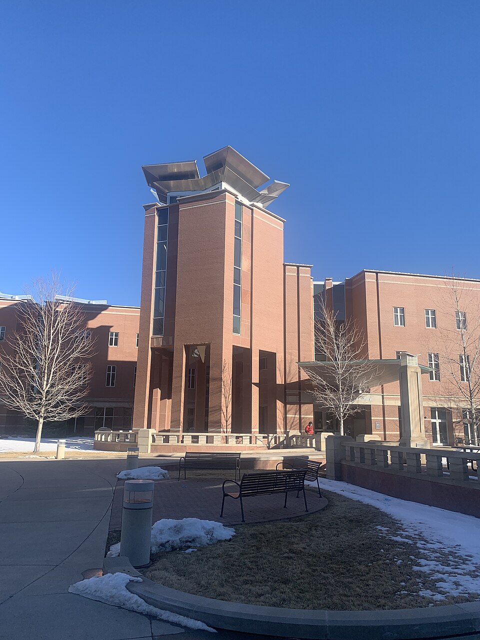 Photograph of the Strauss Health Sciences Library located on the University of Colorado Anschutz Medical Campus in Aurora, Colorado. The image shows the distinctive central tower of the library with its angular metal roof elements under a clear blue sky. Snow is visible on the ground, suggesting the