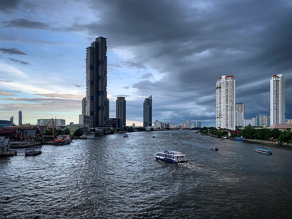 Urban view of the Chao Phraya River in Bangkok, Thailand, featuring several modern skyscrapers along the riverbanks. The buildings include tall, sleek structures with glass facades. The river is dotted with boats, actively navigating its waters. Above, a dramatic cloudy sky looms, creating a strikin