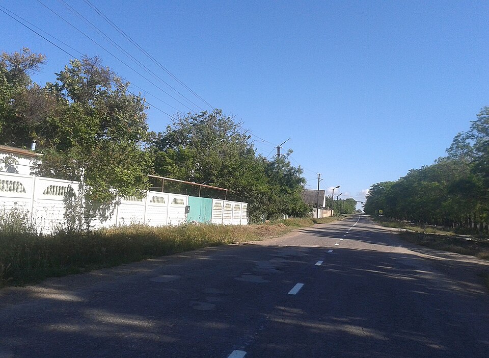 Kirovskoye/Kirovske
Location: Ukraine, Krym
Coordinates: 45.5461643, 33.0883794

Caption: A paved road stretches into the distance under a clear blue sky, flanked by lush green trees and bushes. On the left, a white fence with decorative cutouts runs along the roadside, behind which some overgrown v