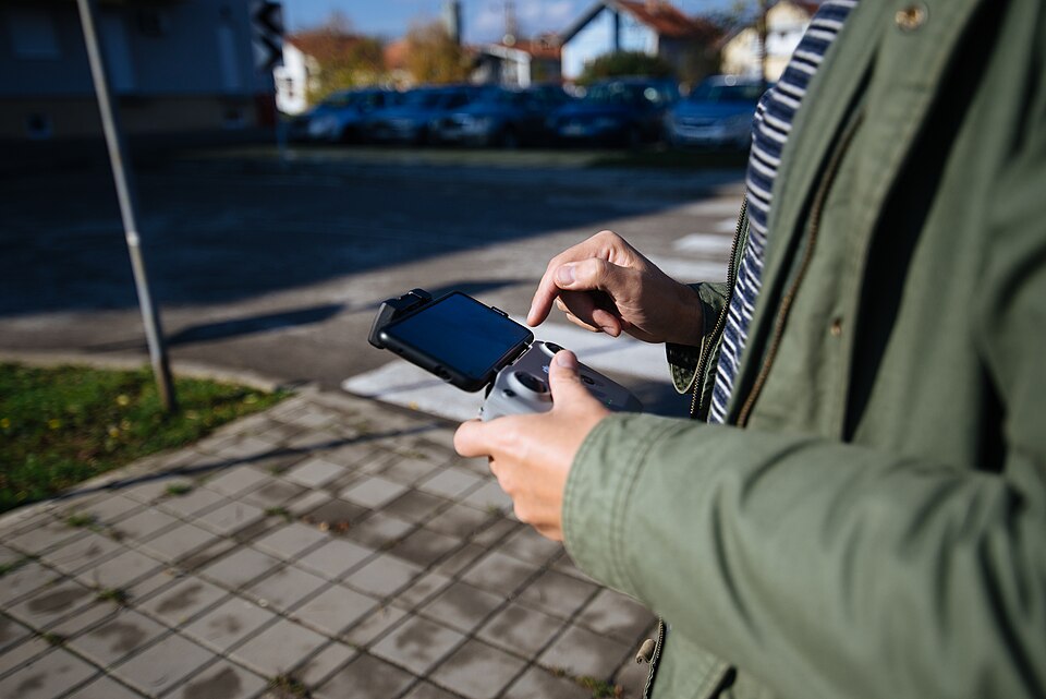 A person stands on a sidewalk, using a drone controller to navigate their device. Surrounding is a quiet suburban neighborhood with parked cars and trees under clear skies.