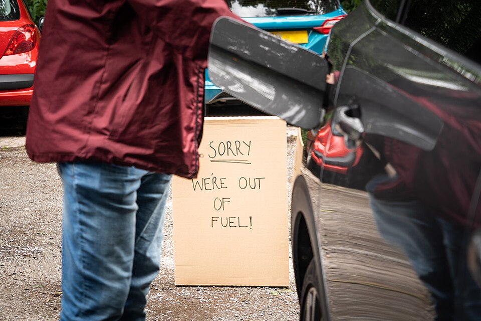 Photograph of a person standing next to a car with the fuel cap open.  There is a sign in the background reading "Sorry, We're out of Fuel!"
This image is free to use on the condition that a link to https://www.icompario.com/nl-be/ is included as attribution.