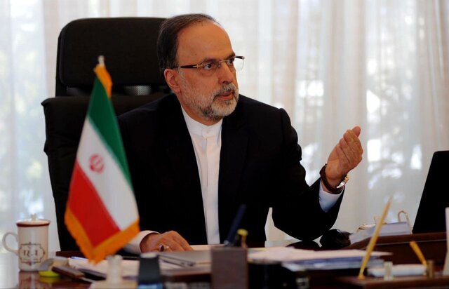 Ahmad Masoumifar, Iranian diplomat and former Ambassador to Sweden and South Korea, seated at his desk during an official meeting or interview. An Iranian flag stands to his left, and various documents, a laptop, and office items are visible on the desk. The photo appears to be taken in an embassy o