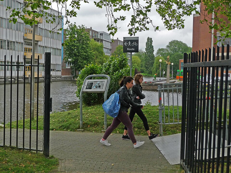 A photo of two students, walking between the fence along the canal Nieuwe Achtergracht on the university campus Roeterseiland, Amsterdam; free photo in high resolutions by Fons Heijnsbroek, June 2013.
