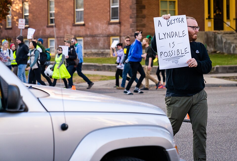Community members gathered at Lyndale Avenue South and West 25th Street in Minneapolis, Minnesota, near where pedestrian Teddy Ferrara had recently been hit and killed by a driver. The group crossed back and forth across Lyndale Avenue, many holding signs encouraging vehicles to slow down and demand
