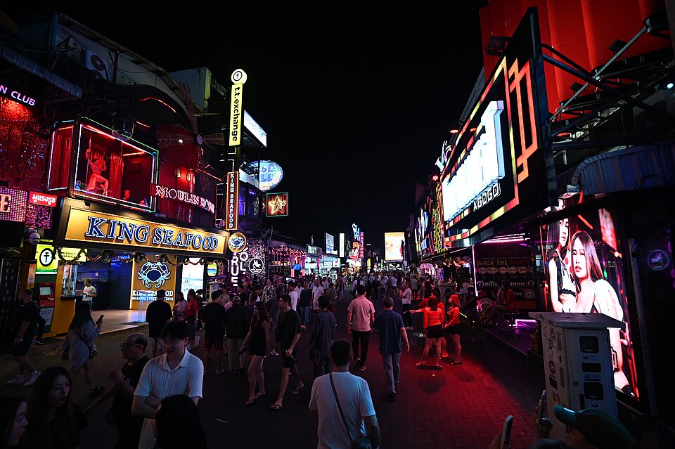 This photo shows a vibrant nightlife scene in what appears to be a bustling entertainment district. The street is illuminated with numerous colorful neon signs and electronic billboards, giving it a distinctive red glow. Several businesses including what looks like "KING OF FOOD" can be seen among t