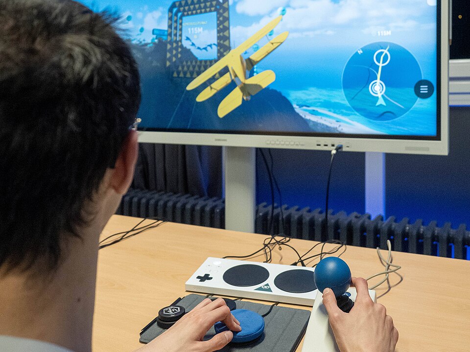 A person sits at a table using the Xbox Adaptive Controller and a large joystick to control a yellow biplane in a flight simulator displayed on a large screen.