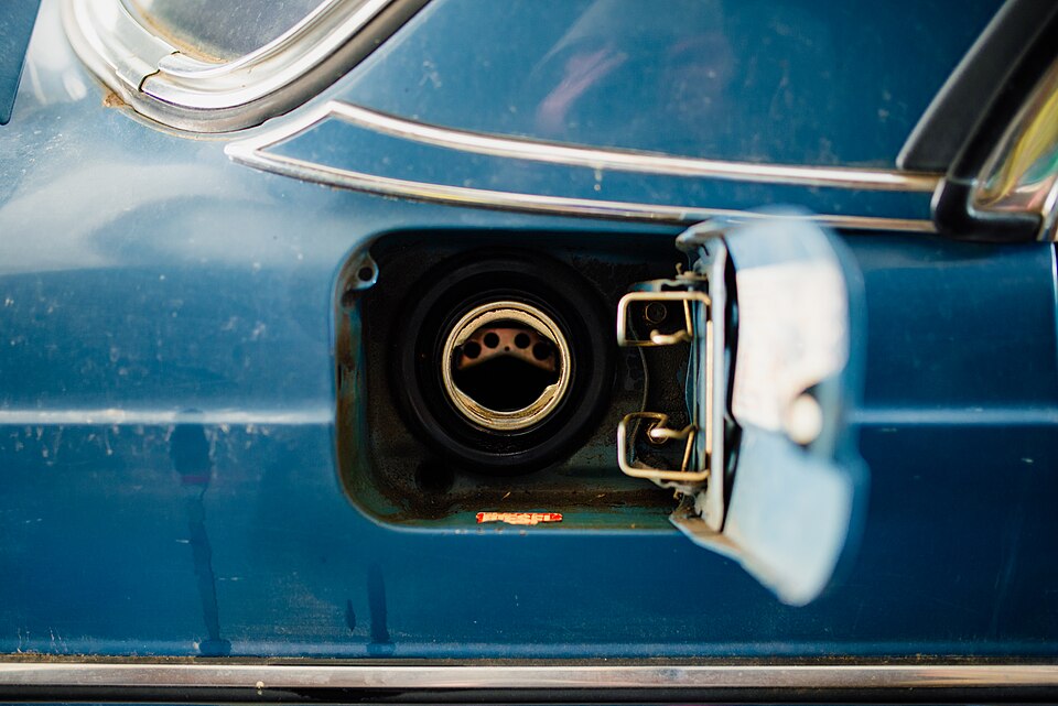 A vintage car's fuel tank opening is revealed at a gas station, showcasing the details of the cap and surrounding area