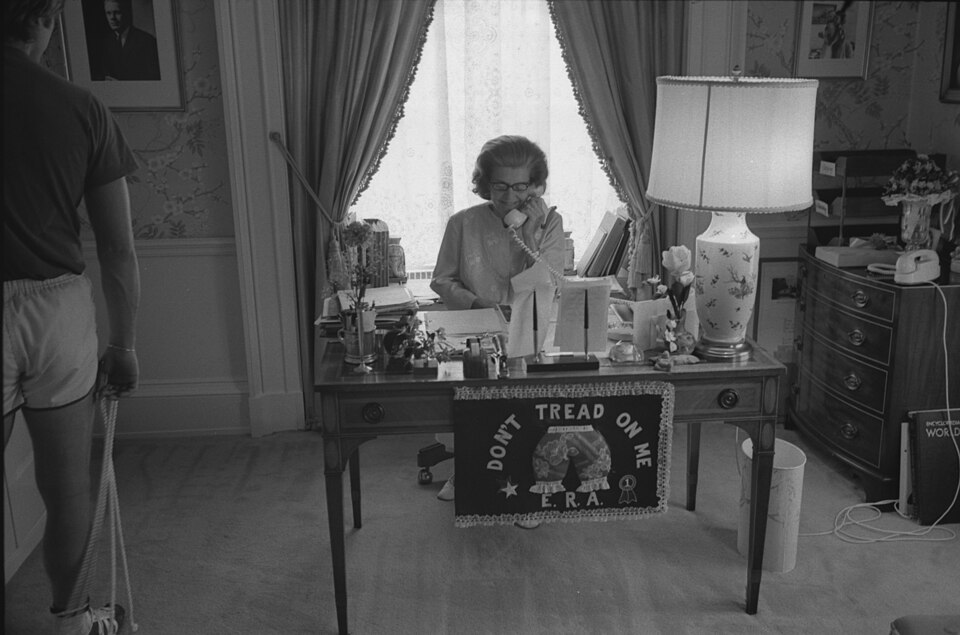 Scope and content:  This photograph depicts First Lady Betty Ford seated at her desk talking on the telephone. Her specially designed "Bloomer" flag showing her support for the Equal Rights Amendment (ERA) is hanging on the front of the desk.