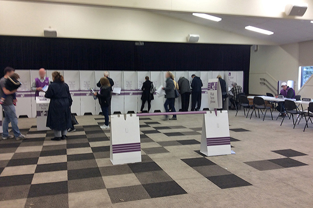 A polling place operating on election day in the 2016 Australian Federal Election. Seated to the right is an electoral official (in purple vest) distributing House of Representatives and Senate ballot papers, voters fill out their papers in polling booths along the wall, while at image left another 
