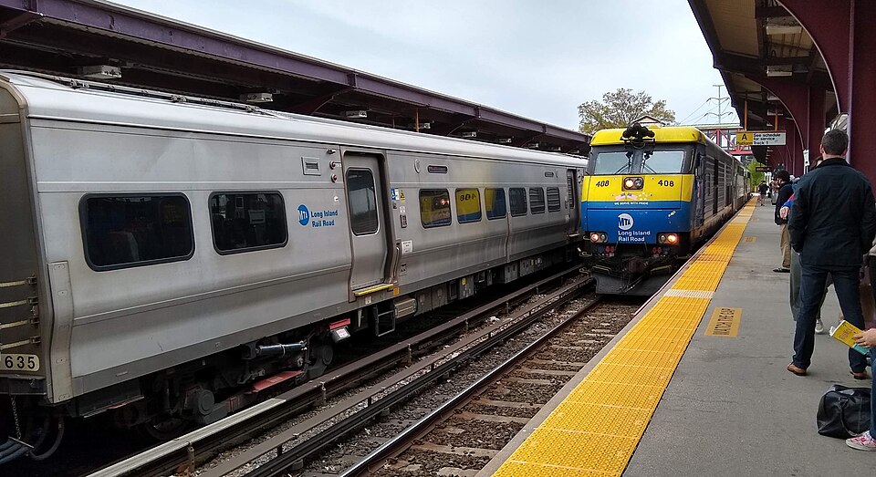Huntington LIRR station on a weekend, taken from Platform A looking west. A diesel train, led by DE30AC #408, is pulling into the station to begin its eastbound run to Port Jefferson and pick up passengers (seen waiting on the platform) from the connecting electric train. An electric train is parked