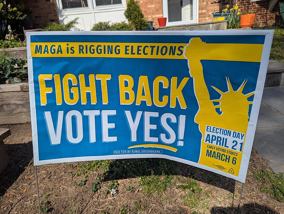Close-up of pro-redistricting amendment yard sign in Virginia, reading "MAGA is Rigging Elections - FIGHT BACK VOTE YES!" with Statue of Liberty silhouette. Paid for by Rural Ground Game. Photographed in Northern Virginia. Election Day April 21, Early Voting starts March 6.