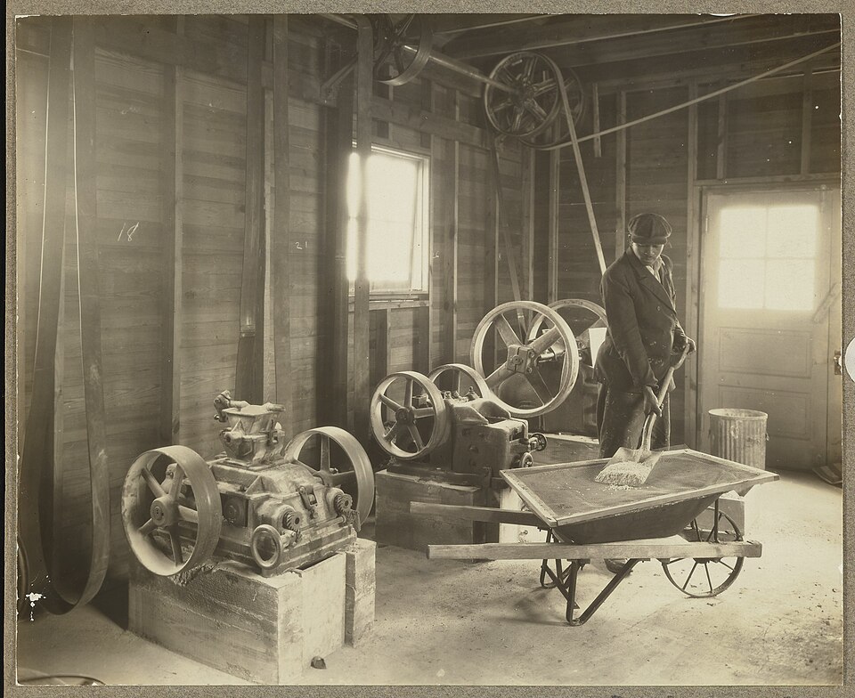 General view of machinery used to crush ore at the Fixed Nitrogen Research Laboratory's blast furnace plant. A worker identified as "Parker" is seen transferring raw material, presumably ore, from a machine into a wheelbarrow for transport. The Fixed Nitrogen Research Laboratory (F.N.R.L.) was estab