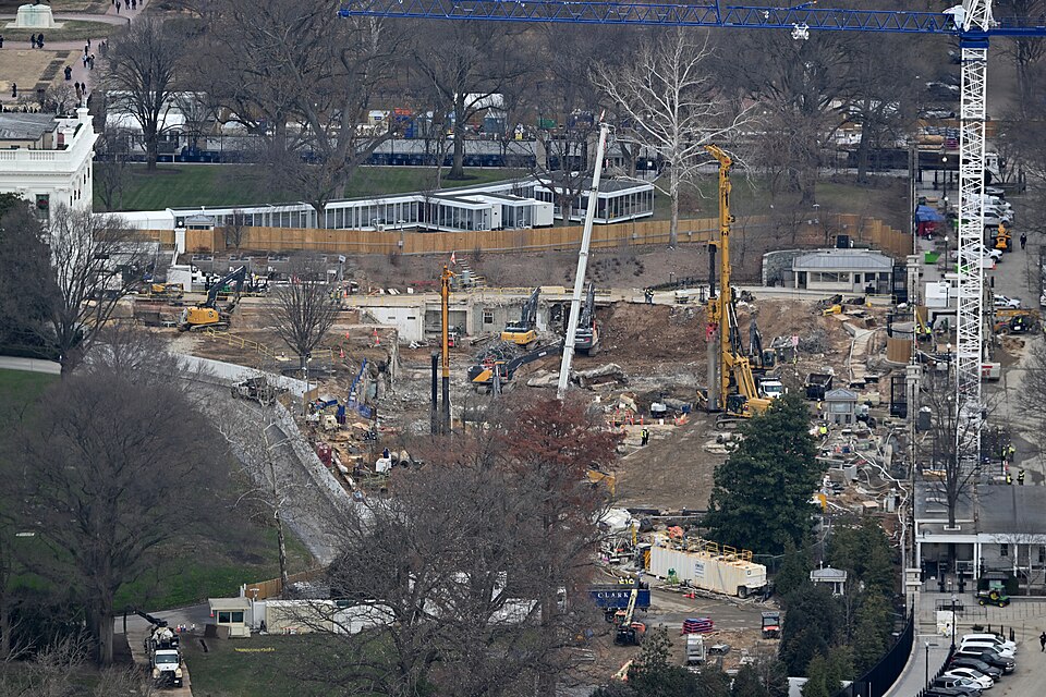 Construction of the White House State Ballroom on December 17, 2025.  The East Wing of the White House has been completely demolished and work at ground level is ongoing.  To the left of the construction site the edge of the White House is visible and behind the construction site is an enclosed walk