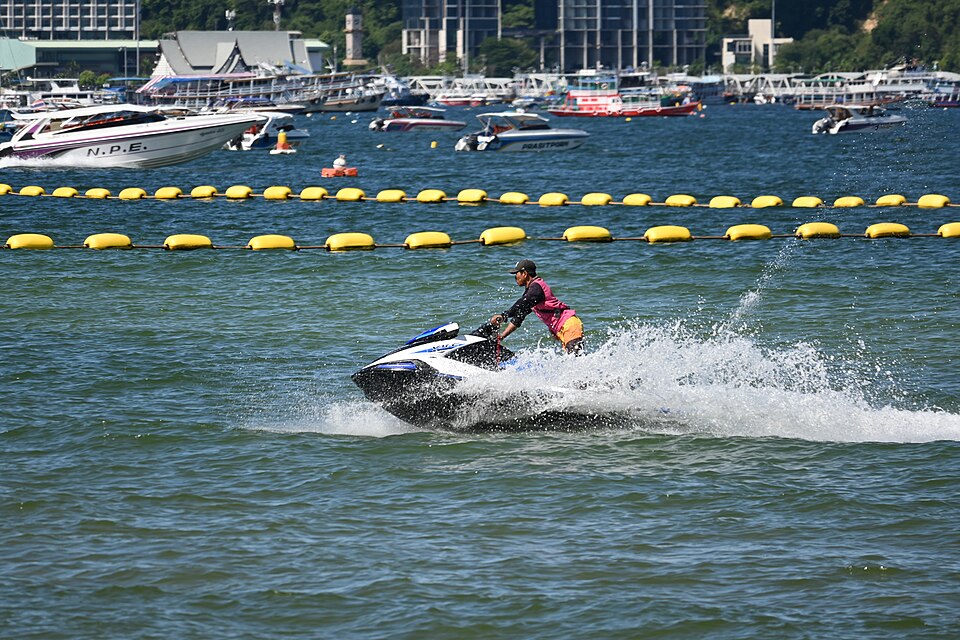 This photo shows a personal watercraft (commonly known as a jet ski) being ridden across a body of water. The rider is creating a spray of water as they speed along. In the background, you can see a line of yellow buoys marking off a swimming or safety area, and beyond that are numerous boats moored